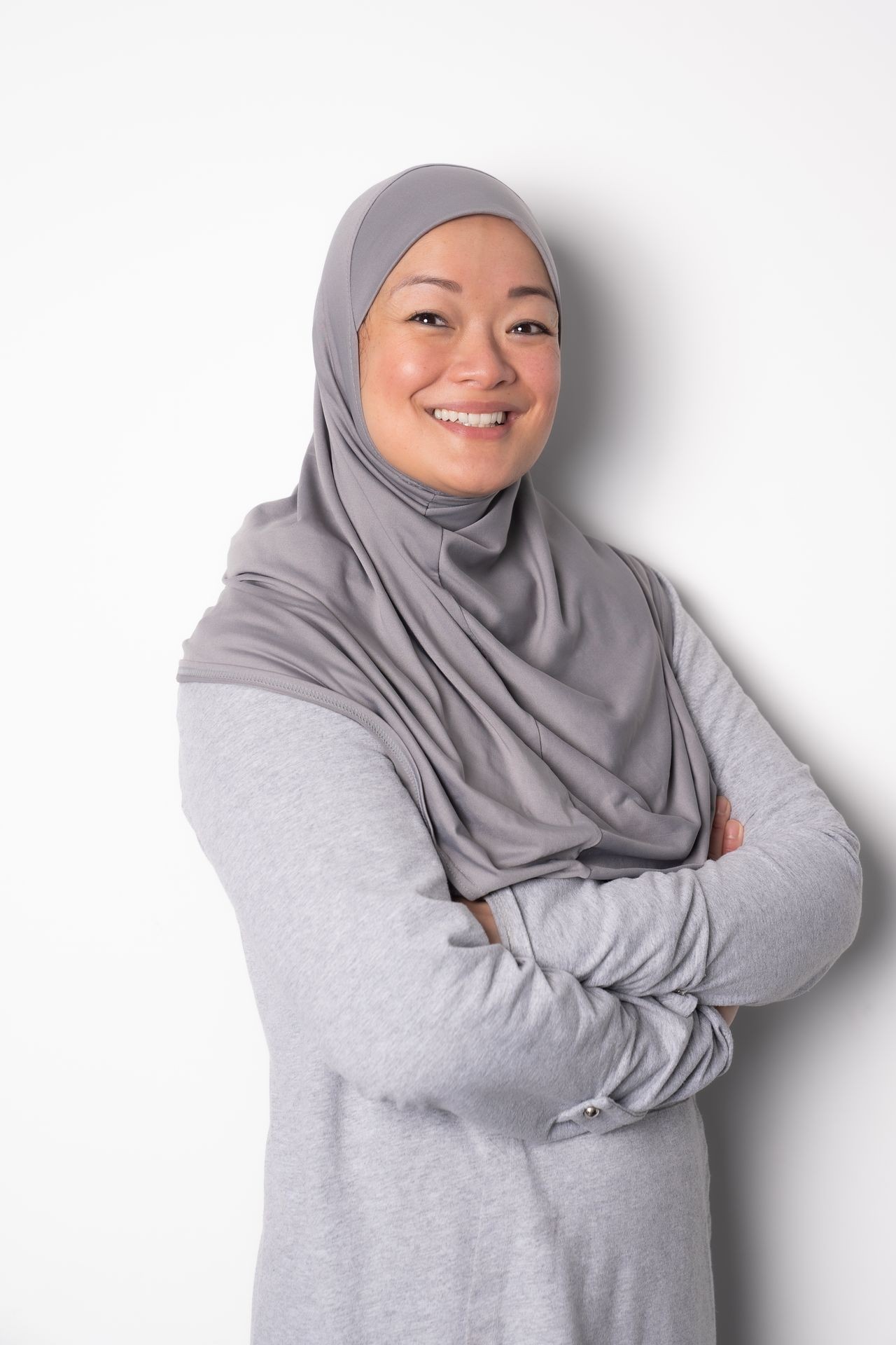 Woman wearing a gray headscarf and sweater, smiling with arms crossed against a white background.