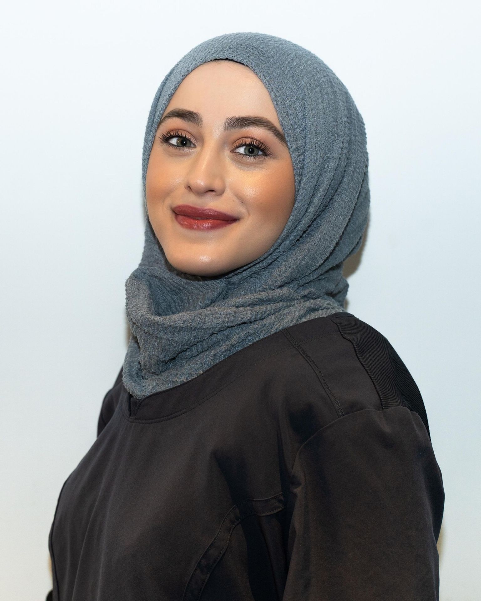 Smiling woman wearing a blue hijab and a dark top against a plain background.