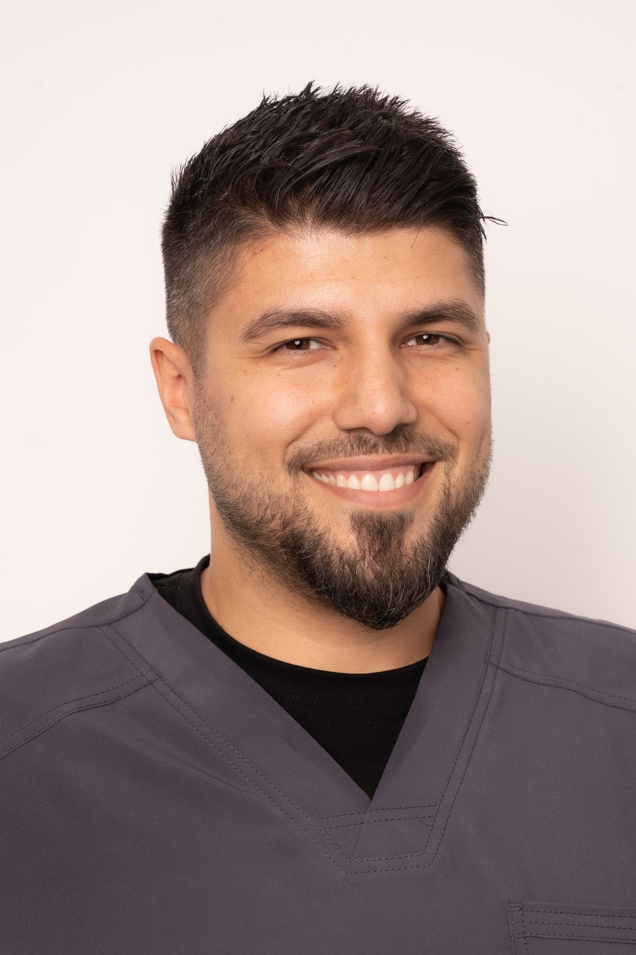 Smiling man wearing a gray V-neck scrub top, against a plain background.