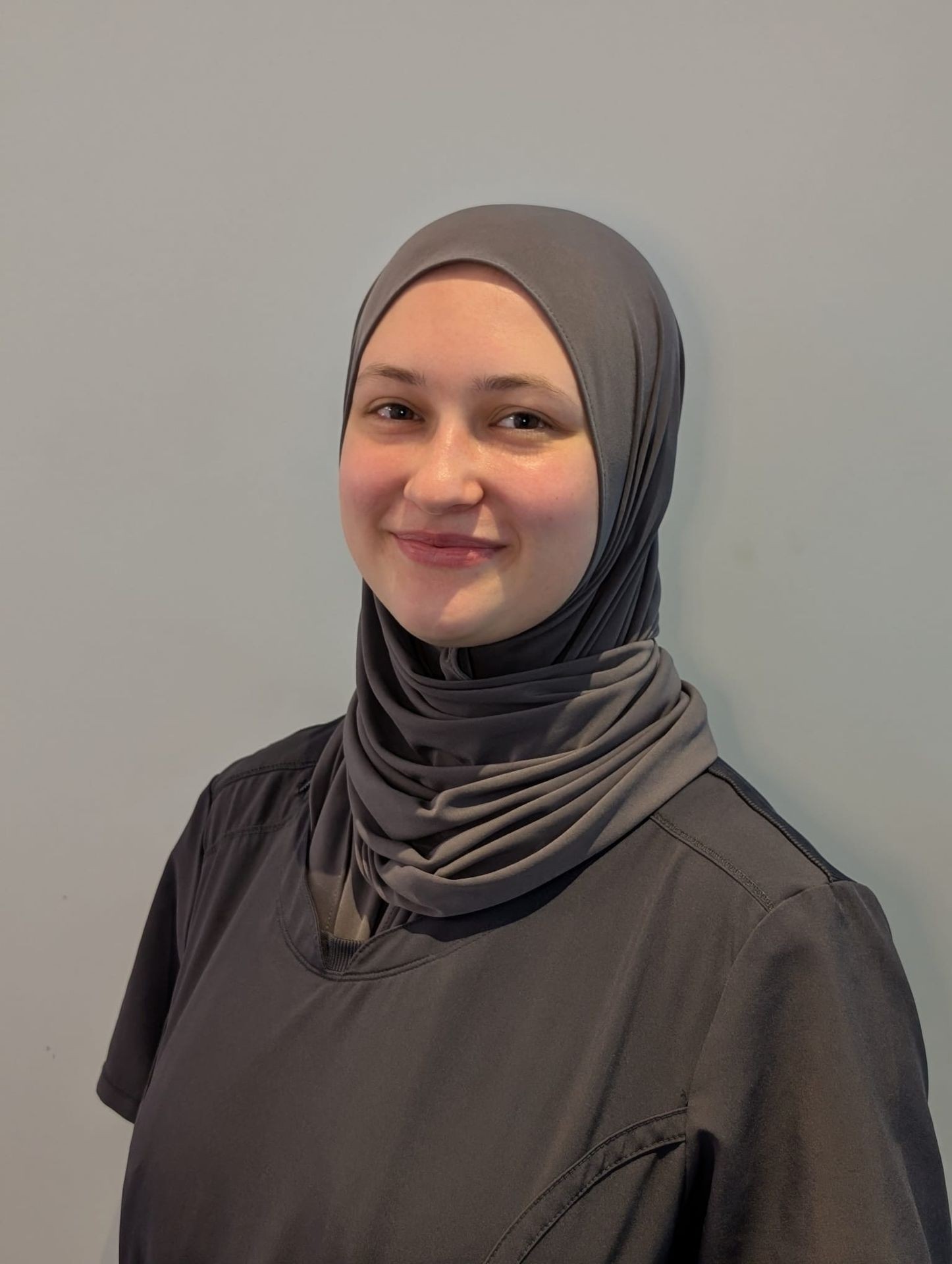 Smiling person wearing a gray hijab and dark clothing against a neutral background.