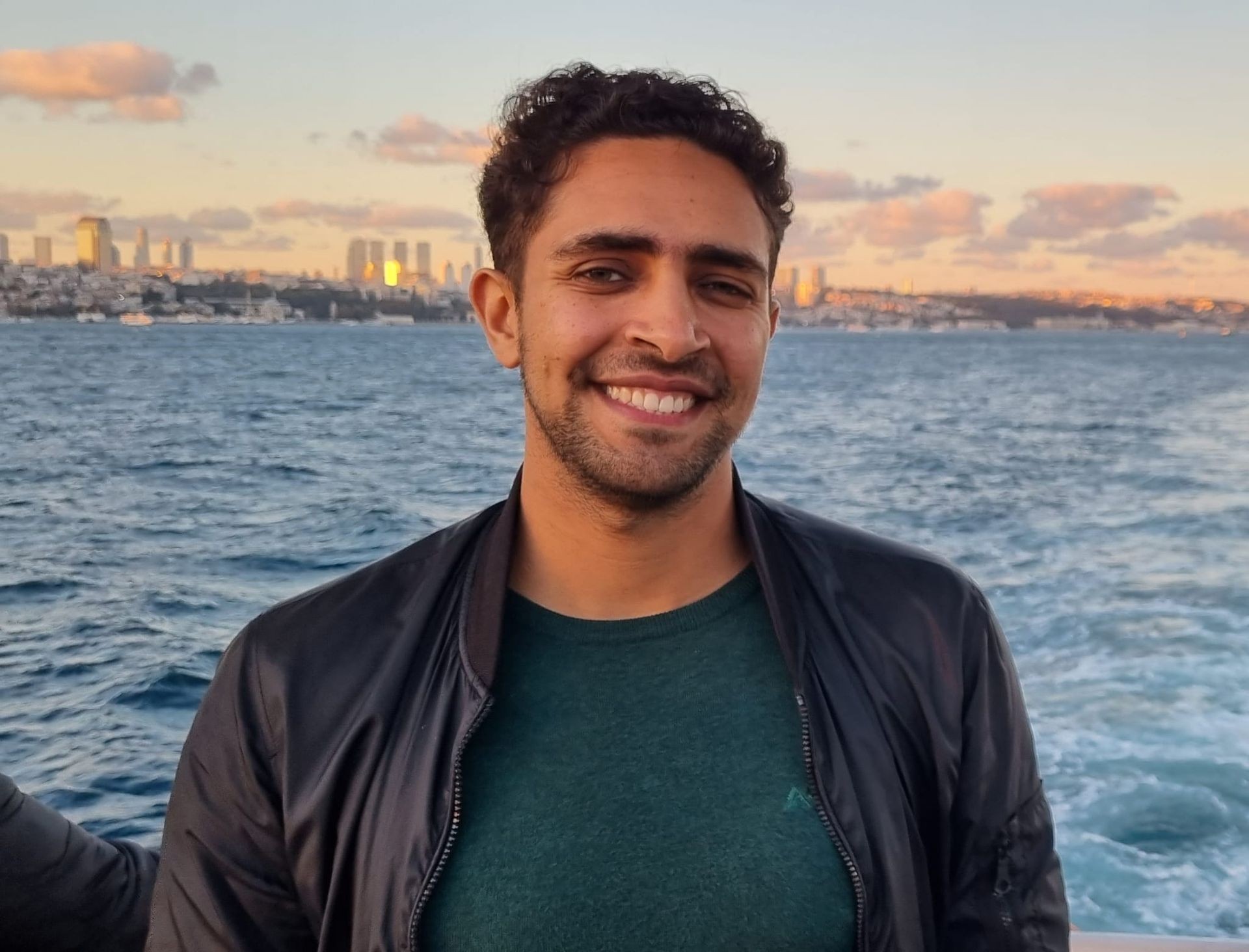 Man smiling in front of a body of water with a city skyline in the background during sunset.